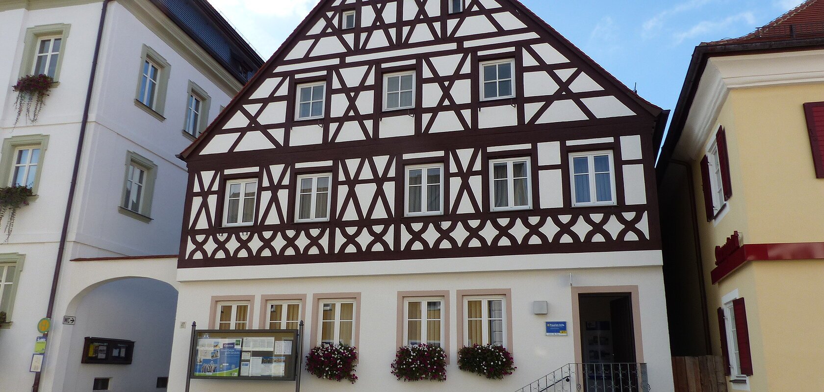 Tourist-Information der Stadt Monheim/Monheimer Alb: ein Fachwerkhaus mit drei Stockwerken, weißen Wänden und braunen Balken. Davor steht eine Sitzbank und ein Schaukasten auf einem gepflasterten Platz. Links davon ein weißes dreistöckiges Gebäude. Rechts davon ein gelbes zweistöckiges Gebäude mit roten Fensterläden.