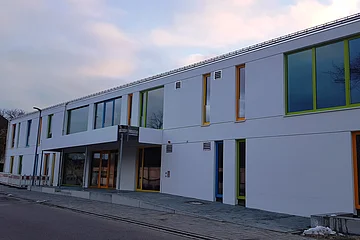 Modernes zweistöckiges Gebäude, der städtische Kindergarten Monheim in weiß, mit vielen bunten Fensterrahmen an einer Straße bei bewölktem Himmel.