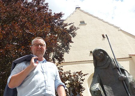 Mann mit Jacke über der Schulter steht neben einer Statue, der Walburga-Statue in Monheim vor einem gelbgestrichenem Gebäude mit Giebeldach. Im Hintergrund ein großer Baum und sonniger Himmel.