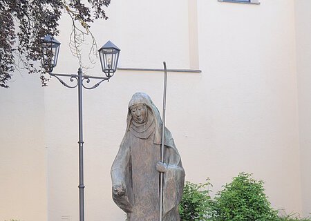 Steinstatue der heiligen Walburga in Monheim mit Stab, umgeben von Blumen in Töpfen und Laternen, vor einer hellen Gebäudewand, der Stadtpfarrkirche Monheim. Vor dem Gebäude sind Pflanzen eingesetzt.