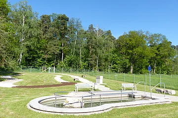 Kneippbecken in Monheim mit Geländer und einem Barfußpfad auf einer Wiese, umgeben von einer großen Grünfläche und Bäumen und Sitzmöglichkeiten. Ein Zaun im Hintergrund.