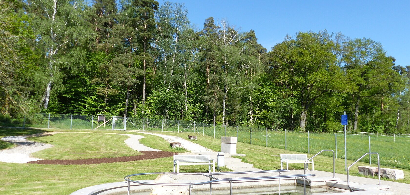 Kneippbecken in Monheim mit Geländer und einem Barfußpfad auf einer Wiese, umgeben von einer großen Grünfläche und Bäumen und Sitzmöglichkeiten. Ein Zaun im Hintergrund.