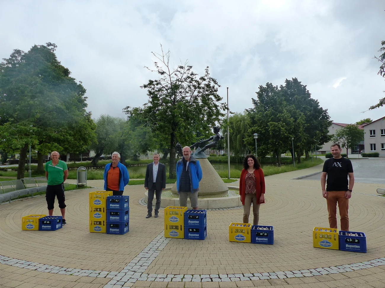 Sechs Personen stehen im Freien auf einem gepflasterten Platz, vor ihnen stehen mehreren Kisten Augustiner-Bier. Im Hintergrund ist ein Stein-Brunnen, Bäume, Gebäude und ein Weiher zu sehen.