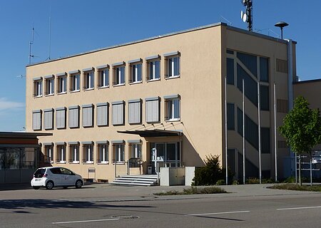 Dreistöckiges Gebäude mit beigen Wänden, die Caritas Sozial-Station in Monheim, mit vielen Fenstern, einer Treppe vor dem Gebäude und einem geparkten Auto auf dem Parkplatz. Vorderhalb des Gebäudes verläuft eine Straße.
