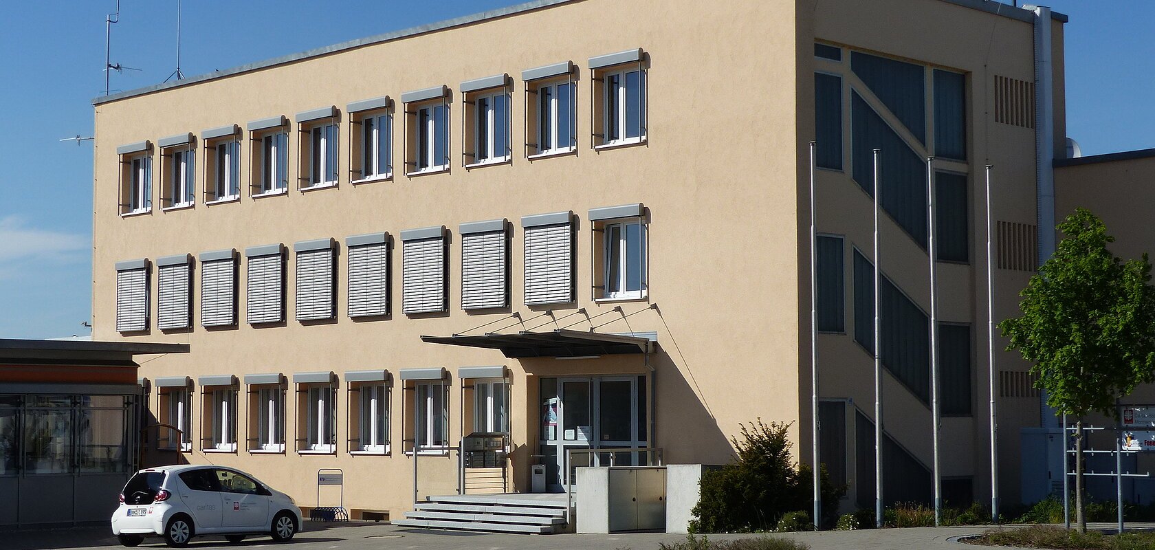 Dreistöckiges Gebäude mit beigen Wänden, die Caritas Sozial-Station in Monheim, mit vielen Fenstern, einer Treppe vor dem Gebäude und einem geparkten Auto auf dem Parkplatz. Vorderhalb des Gebäudes verläuft eine Straße.