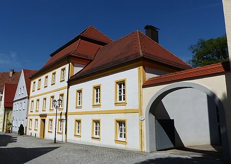 Historisches weißes Gebäude, die Pfarr- und Stadtbücherei in Monheim, mit gelben Fensterrahmen und rotem Dach, daneben ein Torbogen auf gepflasterter Straße. Links vom Gebäude weitere Häuser in blau und grün.