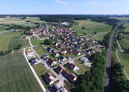 Luftaufnahme des Dorfes Weilheim, mit vielen Häusern, umgeben von Feldern und Wäldern, neben einer Bahnstrecke.