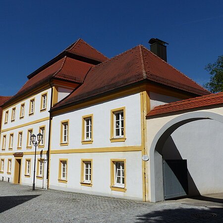 Historisches weißes Gebäude, die Pfarr- und Stadtbücherei in Monheim, mit gelben Fensterrahmen und rotem Dach, daneben ein Torbogen auf gepflasterter Straße. Links vom Gebäude weitere Häuser in blau und grün.
