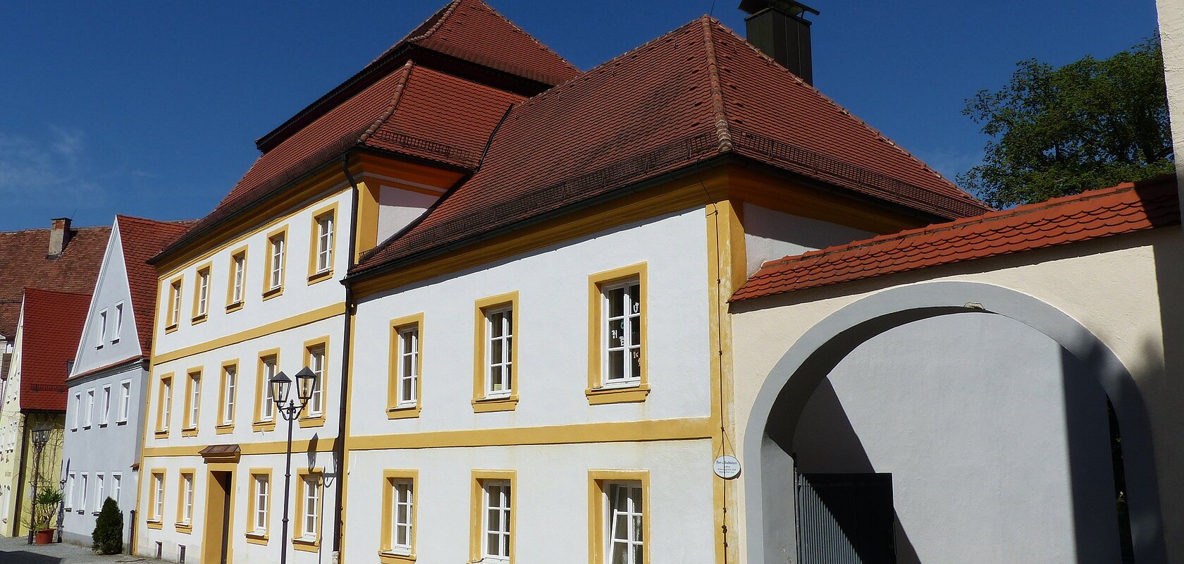 Historisches weißes Gebäude, die Pfarr- und Stadtbücherei in Monheim, mit gelben Fensterrahmen und rotem Dach, daneben ein Torbogen auf gepflasterter Straße. Links vom Gebäude weitere Häuser in blau und grün.