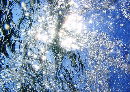 Lichtreflexionen und Luftblasen unter Wasser, mit blauem Himmel über dem Wasser.