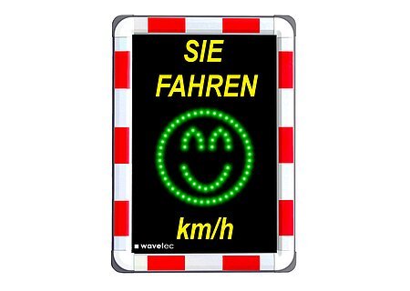 Bild eines Geschwindigkeitsmessgeräts im Straßenverkehr, darauf oben gelber Text "Sie fahren" und unten "km/h". Dazwischen ein grünes Smiley-Gesicht aus LEDs. Der Rand außen ist rot/weiß.