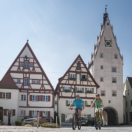 Zwei Personen fahren mit Fahrrädern auf einem gepflasterten Platz, der Monheimer Altstadt. Im Hintergrund Fachwerkhäuser und ein Turm mit einem Torbogen und einer Uhr.