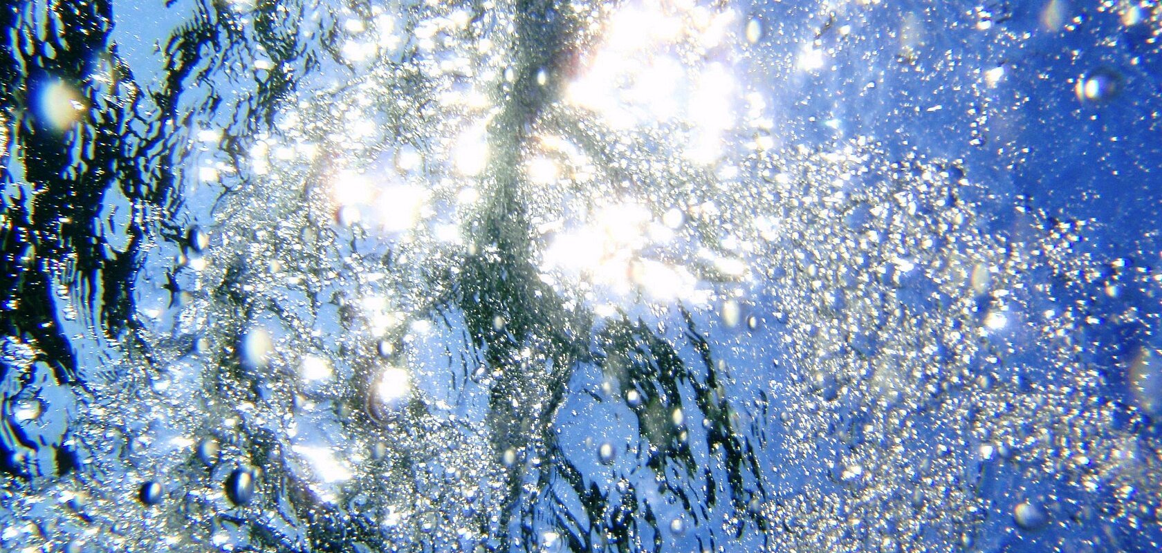Lichtreflexionen und Luftblasen unter Wasser, mit blauem Himmel über dem Wasser.