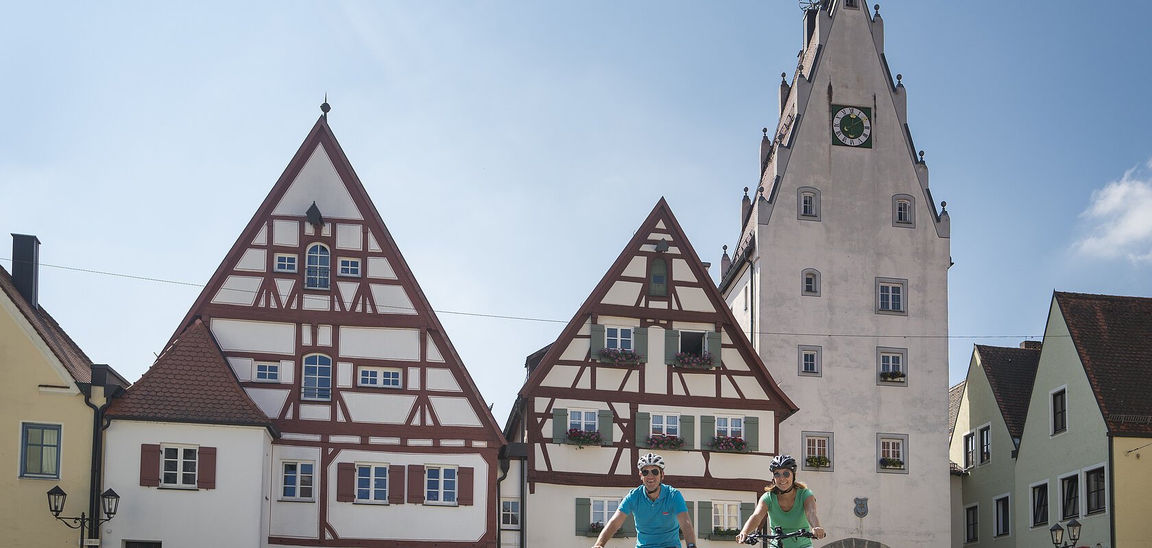 Zwei Personen fahren mit Fahrrädern auf einem gepflasterten Platz, der Monheimer Altstadt. Im Hintergrund Fachwerkhäuser und ein Turm mit einem Torbogen und einer Uhr.