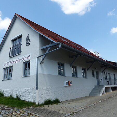 Weißes Gebäude mit rotem Dach, einer Treppe und einem Defibrillator. steht auf einem gepflasterten Grundstück. Auf dem Gebäude der Schriftzug "FFW Flotzheim-Kreut" an der Fassade. Blauer Himmel und Bäume im Hintergrund.