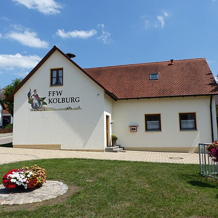 Ein gelbes Gebäude, das Feuerwehrhaus in Kölburg mit rotem Dach, vier Fenstern und einem Defibrillator. Auf dem Gebäude die Aufschrift "FFW Kolburg", davor ein Blumenbeet auf einer Rasenfläche und ein weiterer Blumenkasten auf einem Geländer. Im Hintergrund eine Straße mit einem weißen Haus.