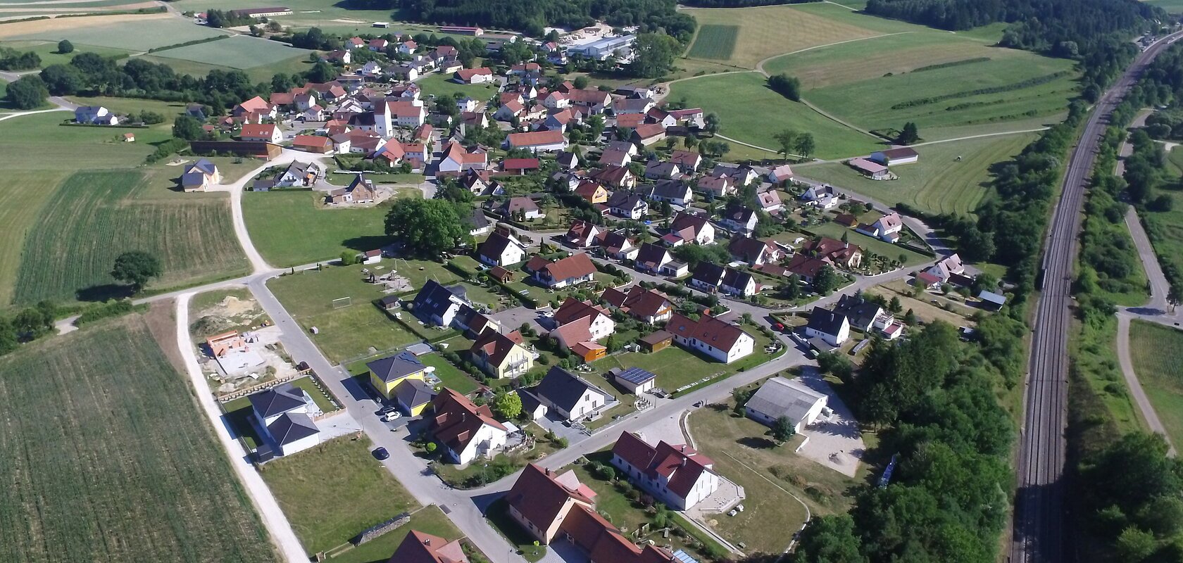 Luftaufnahme des Dorfes Weilheim, mit vielen Häusern, umgeben von Feldern und Wäldern, neben einer Bahnstrecke.