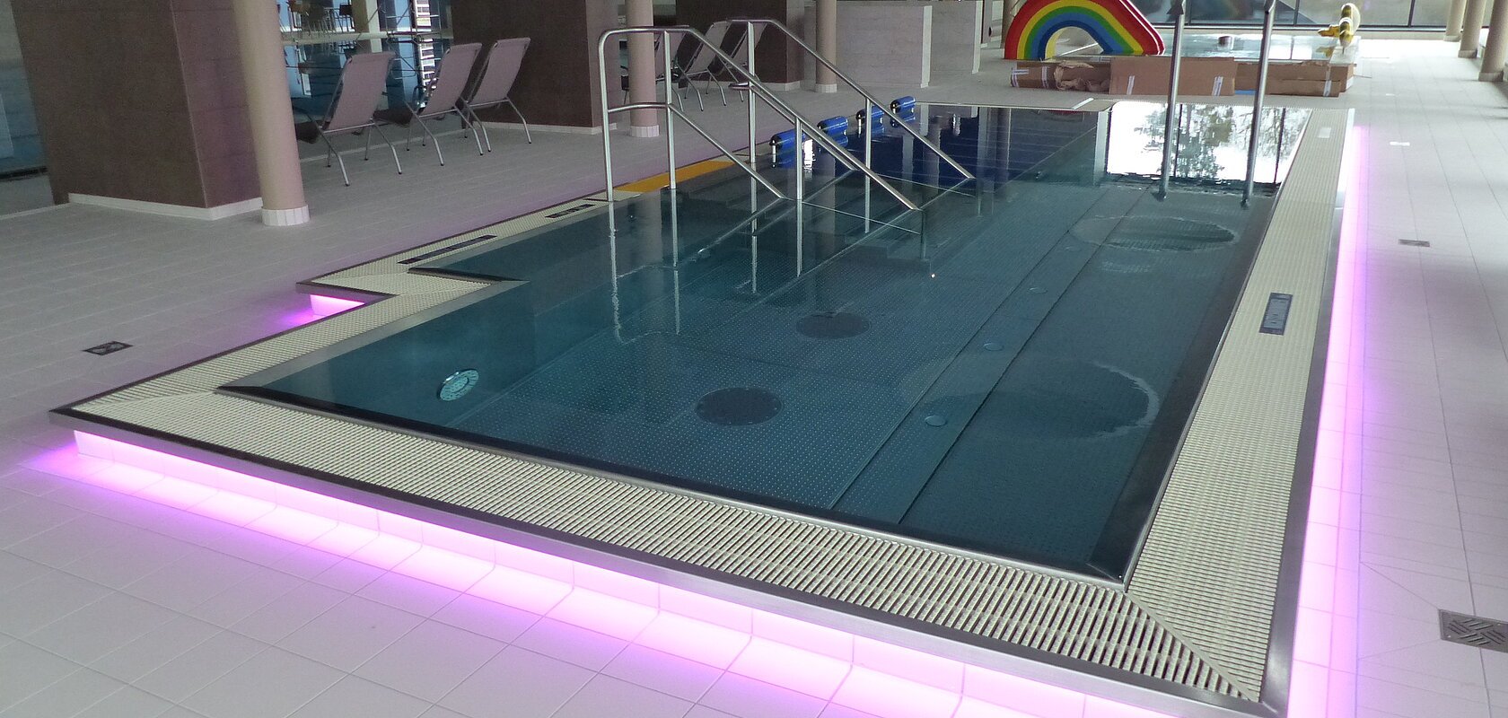 Innenansicht eines Schwimmbades, dem Monheimer Jurabad: ein mit Wasser befülltes Schwimmbecken mit Stufen und Handläufen, umgeben von Fliesenboden. Der Rand des Beckens ist außenrum rosa/lila beleuchtet. Im Hintergrund stehen drei Liegestühle und eine Regenbogenrutsche, sowie ein weiteres Becken.