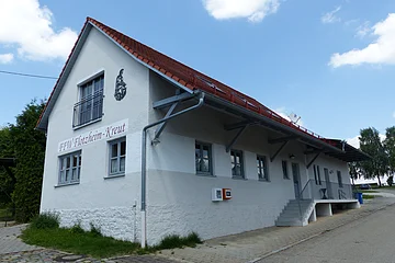 Weißes Gebäude mit rotem Dach, einer Treppe und einem Defibrillator. steht auf einem gepflasterten Grundstück. Auf dem Gebäude der Schriftzug "FFW Flotzheim-Kreut" an der Fassade. Blauer Himmel und Bäume im Hintergrund.