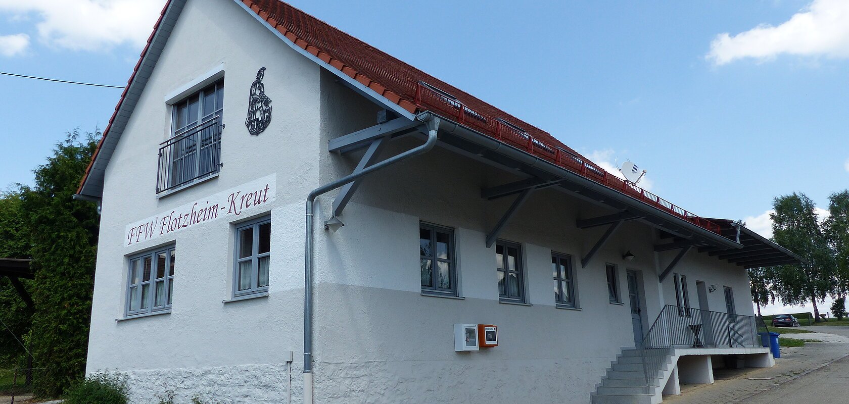 Weißes Gebäude mit rotem Dach, einer Treppe und einem Defibrillator. steht auf einem gepflasterten Grundstück. Auf dem Gebäude der Schriftzug "FFW Flotzheim-Kreut" an der Fassade. Blauer Himmel und Bäume im Hintergrund.