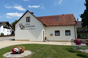 Ein gelbes Gebäude, das Feuerwehrhaus in Kölburg mit rotem Dach, vier Fenstern und einem Defibrillator. Auf dem Gebäude die Aufschrift "FFW Kolburg", davor ein Blumenbeet auf einer Rasenfläche und ein weiterer Blumenkasten auf einem Geländer. Im Hintergrund eine Straße mit einem weißen Haus.