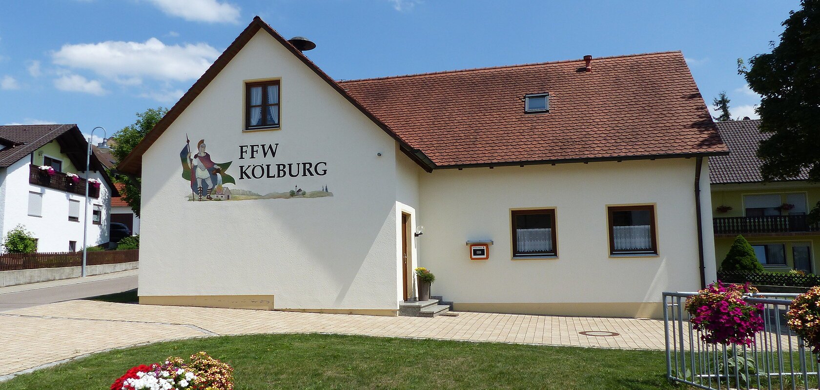 Ein gelbes Gebäude, das Feuerwehrhaus in Kölburg mit rotem Dach, vier Fenstern und einem Defibrillator. Auf dem Gebäude die Aufschrift "FFW Kolburg", davor ein Blumenbeet auf einer Rasenfläche und ein weiterer Blumenkasten auf einem Geländer. Im Hintergrund eine Straße mit einem weißen Haus.