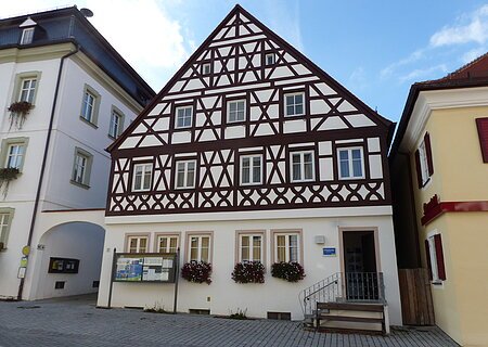 Tourist-Information der Stadt Monheim/Monheimer Alb: ein Fachwerkhaus mit drei Stockwerken, weißen Wänden und braunen Balken. Davor steht eine Sitzbank und ein Schaukasten auf einem gepflasterten Platz. Links davon ein weißes dreistöckiges Gebäude. Rechts davon ein gelbes zweistöckiges Gebäude mit roten Fensterläden.
