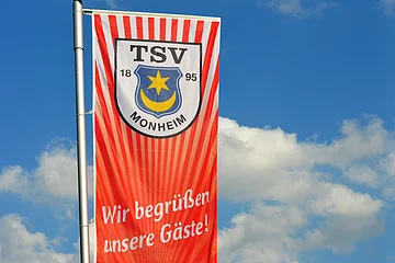 TSV 1895 Monheim e.V. - Wir begrüßen unsere Gäste! Rote Fahne mit dem Logo des TSV Monheim und dem Text "Wir begrüßen unsere Gäste!" vor blauem Himmel.