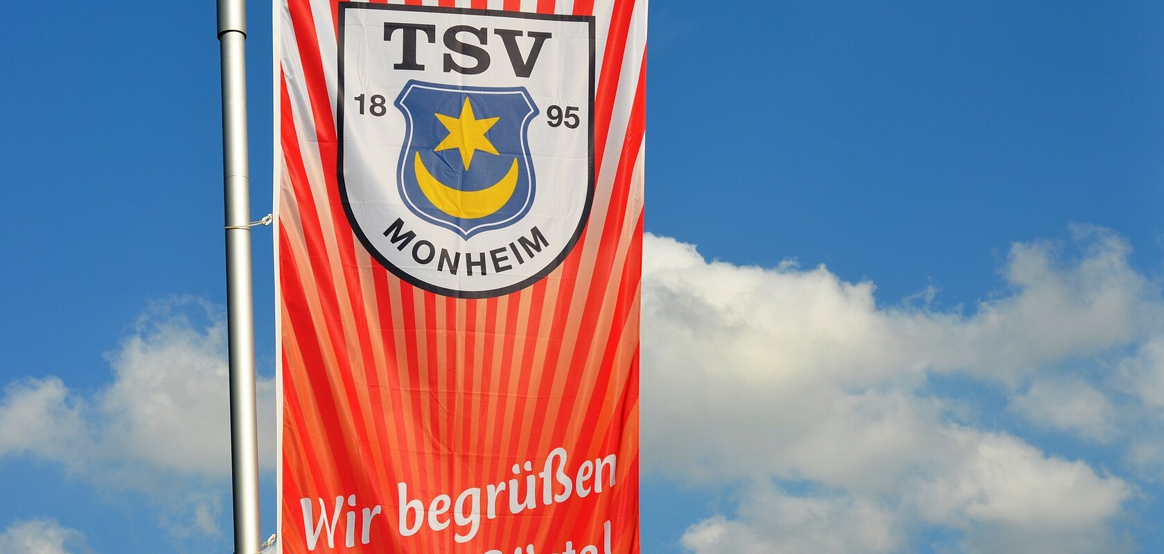 TSV 1895 Monheim e.V. - Wir begrüßen unsere Gäste! Rote Fahne mit dem Logo des TSV Monheim und dem Text "Wir begrüßen unsere Gäste!" vor blauem Himmel.