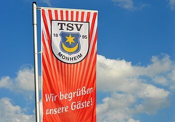 Rote Fahne mit dem Logo des TSV Monheim und dem Text "Wir begrüßen unsere Gäste!" vor blauem Himmel.