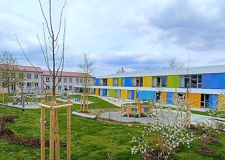Zwei Gebäude des städtischen Kindergartens Monheim, eins davon mit einer bunten Fassade, davor ein Garten mit Spielgeräten Bäumen und Rasenfläche, bewölkter Himmel.