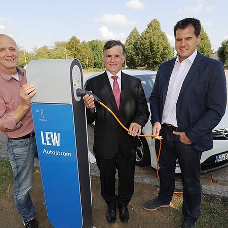 Drei Männer stehen neben einer LEW Autostrom-Ladestation mit dem Kabel in der Hand. Hinter Ihnen ein weißes Elektroauto auf einem Parkplatz. Im Hintergrund sind Bäume zu sehen.