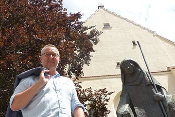 Mann mit Jacke über der Schulter steht neben einer Statue, der Walburga-Statue in Monheim vor einem gelbgestrichenem Gebäude mit Giebeldach. Im Hintergrund ein großer Baum und sonniger Himmel.