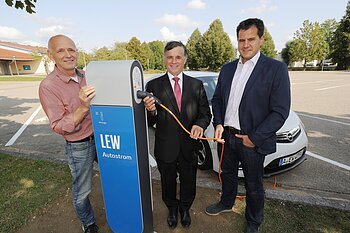E-Car Ladestation in Monheim (auf dem Bild: LEW-Kommunalbetreuer Ulrich Endraß, Günther Pfefferer, Erster Bürgermeister von Monheim, und Peter Ferber, Stadt-Aktiv-Manager in Monheim bei der Inbetriebnahme der Ladestation) Drei Männer stehen neben einer LEW Autostrom-Ladestation mit dem Kabel in der Hand. Hinter Ihnen ein weißes Elektroauto auf einem Parkplatz. Im Hintergrund sind Bäume zu sehen.