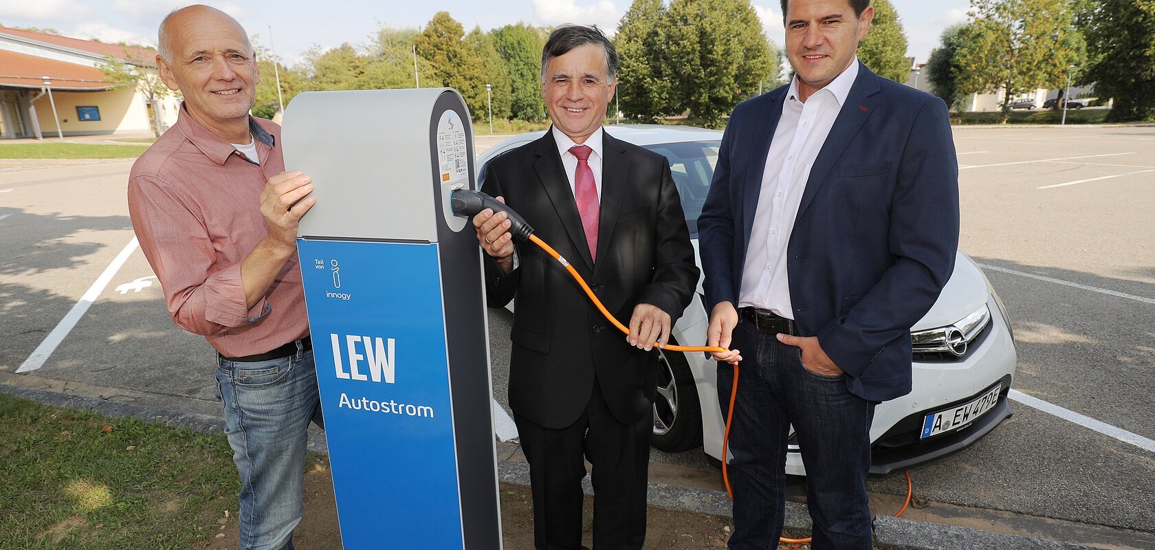 Drei Männer stehen neben einer LEW Autostrom-Ladestation mit dem Kabel in der Hand. Hinter Ihnen ein weißes Elektroauto auf einem Parkplatz. Im Hintergrund sind Bäume zu sehen.