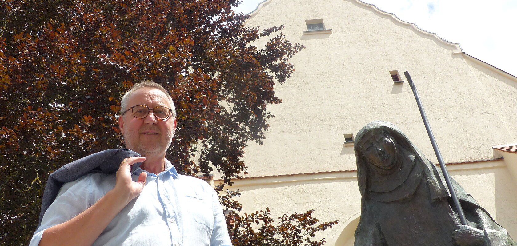 Mann mit Jacke über der Schulter steht neben einer Statue, der Walburga-Statue in Monheim vor einem gelbgestrichenem Gebäude mit Giebeldach. Im Hintergrund ein großer Baum und sonniger Himmel.