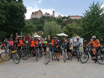 Gruppe von Radfahrern teilweise in orangefarbenen Trikots posiert mit Fahrrädern vor einer Burg, der Harburg auf einem gepflasterten Platz. Im Hintergrund unterhalb der Burg sind Bäume.