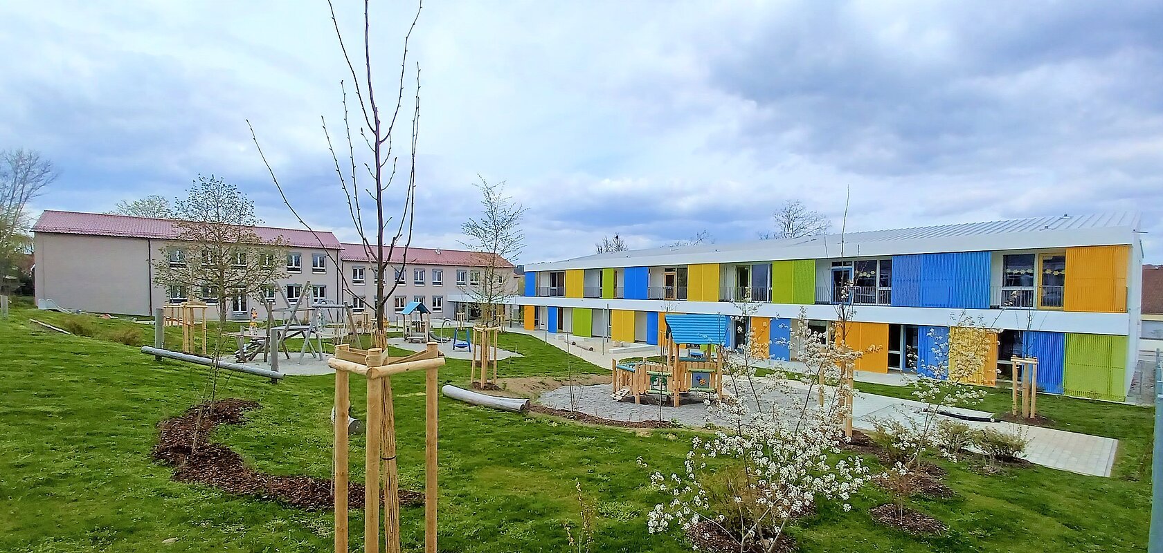 Zwei Gebäude des städtischen Kindergartens Monheim, eins davon mit einer bunten Fassade, davor ein Garten mit Spielgeräten Bäumen und Rasenfläche, bewölkter Himmel.