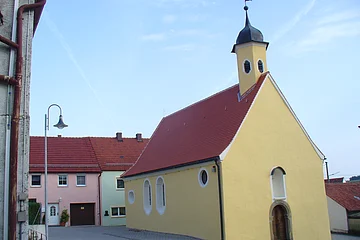 Peterskapelle Monheim Gelbe Kapelle, die Peterskapelle in Monheim mit rotem Dach und einem Turm, umgeben von Wohnhäusern mit roten Dächern. Im Hintergrund blauer Himmel.