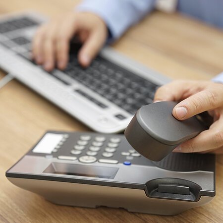 Person im blauen Hemd tippt auf einer Computertastatur und hält den Hörer eines Tischtelefons in der Hand.