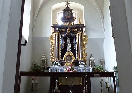 Ein Blick durch eine Tür zu einem Kirchenaltar in der Walburgakapelle in Monheim, mit goldenen Verzierungen und einer Statue, der heiligen Walburga. Der Kirchenaltar ist umgeben von Kerzen und Blumen, in einem gewölbten Raum mit einer verzierten Decke.