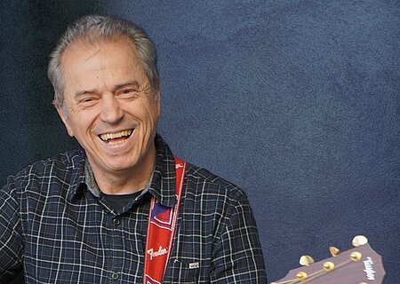 Ein älterer Mann spielt lächelnd Akustikgitarre vor einem blauen Hintergrund.