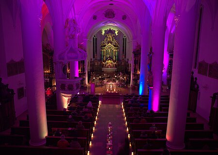 Innenansicht einer Kirche mit violetter Beleuchtung, Kerzen entlang des Gangs und Menschen auf den Bänken. Vor dem Altar spielt eine Musik-Kapelle.