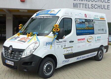 Ein weißer Transporter vom Car-Sharing-Anbieter Mikar mit Werbeaufdrucken und Sonnenblumen steht vor einem Gebäude mit der Aufschrift "Grundschule Mittelschule" in Monheim.