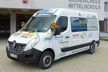Ein weißer Transporter vom Car-Sharing-Anbieter Mikar mit Werbeaufdrucken und Sonnenblumen steht vor einem Gebäude mit der Aufschrift "Grundschule Mittelschule" in Monheim.