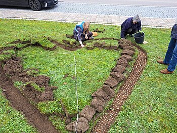 "Monheim summt!" - Herz 2023 Zwei Personen pflanzen Blumenzwiebeln in ein herzförmig angelegtes Beet auf einer Rasenfläche. Erde ist auf beiden Seiten aufgeschichtet. Rechts die Beine einer weiteren Person zu sehen. Vor dem Rasen ein gepflasterter Fußweg und eine Straße mit einem parkendem Auto drauf.