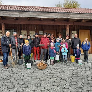 Zwiebelpflanzaktion 2022 in Monheim - 15.500 Blumenzwiebeln - 4 Stunden und viele helfende Hände Gruppe von Menschen mit Gartenwerkzeugen und Eimern und einem geflochtenen Korb, steht vor einem Holzgebäude auf gepflastertem Boden.