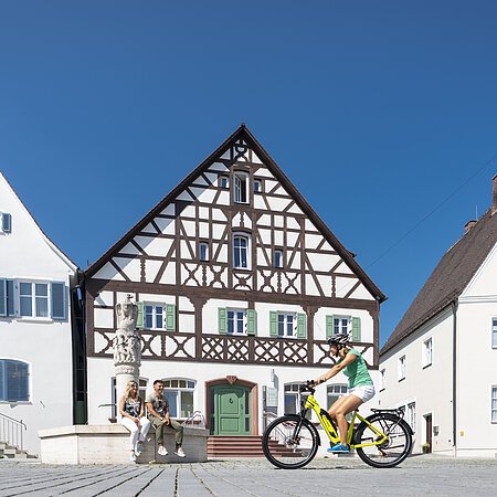 Der Marktplatz in Monheim: ein Fachwerkhaus zwischen zwei Gebäuden, ein Fahrradfahrer im Vordergrund fährt auf dem gepflastertem Platz, zwei Personen sitzen auf dem Rand eines Brunnens vor dem Fachwerkhaus. Auf dem linken Gebäude steht die Aufschrift "Stadtapotheke", auf dem rechten Gebäude die Aufschrift "Sparkasse".