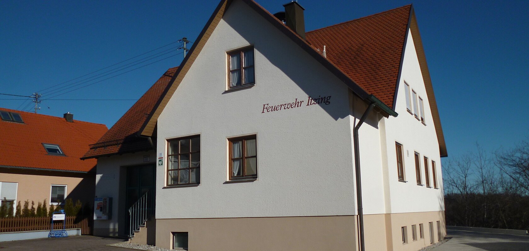 Weißes Feuerwehrgebäude mit rotem Dach, Aufschrift "Feuerwehr Izing" an der Fassade, gepflasterter Vorplatz, blauer Himmel. Links davon ein weiteres Haus in orange/beige mit rotem Dach. Vorderhalb gepflasterter Platz und blauer Himmel im Hintergrund.