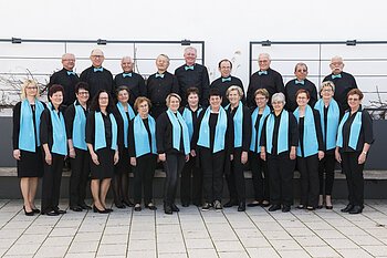 Gruppe von 21 Personen, dem Liederkranz Monheim, in schwarzer Kleidung mit türkisfarbenen Schals und Fliegen, posiert vor einer weißen Wand auf einem gepflastertem Platz.